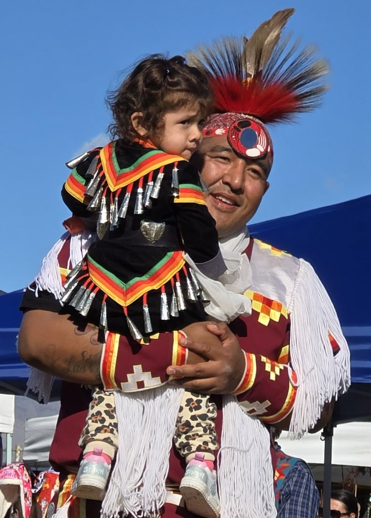 A happy Native American father holding daughter in Jingle Dress as seen in powwows: Cedar Band; Cedar City; Duck Valley; Elko; Ely; Fallon; Fort Mojave; Goshute; Las Vegas; Snow Mountain; Las Vegas Snow Mountain; Las Vegas Paiute Tribe; Las Vegas Snow Mountain Paiute; Las Vegas Snow Mountain Paiute Tribe; 33rd annual Las Vegas Snow Mountain Paiute; Moapa; Muckleshoot; Nevada Day; Numaga; Pahrump; Pahrump Valley; Pahrump Valley Social; Pahrump Social; Petrack; Petrack Park; Red Dress; Sacred Visions; San Marcos; San Marcos Apache; Stewart; Sycuan; UNLV; UNR; Univ. of Nev. Reno; and Walker River; & Western Navajo Fair.